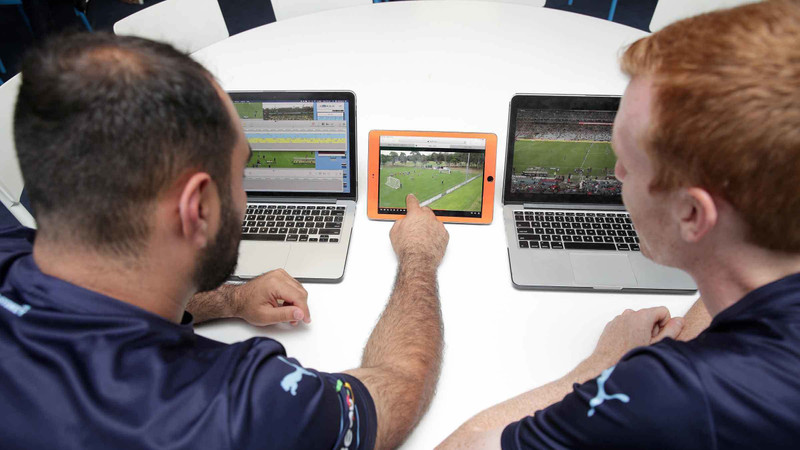 2 台のノート PC と 1 台のタブレットでサッカーのビデオ映像の分析を見ている 2 人のシドニー FC 選手